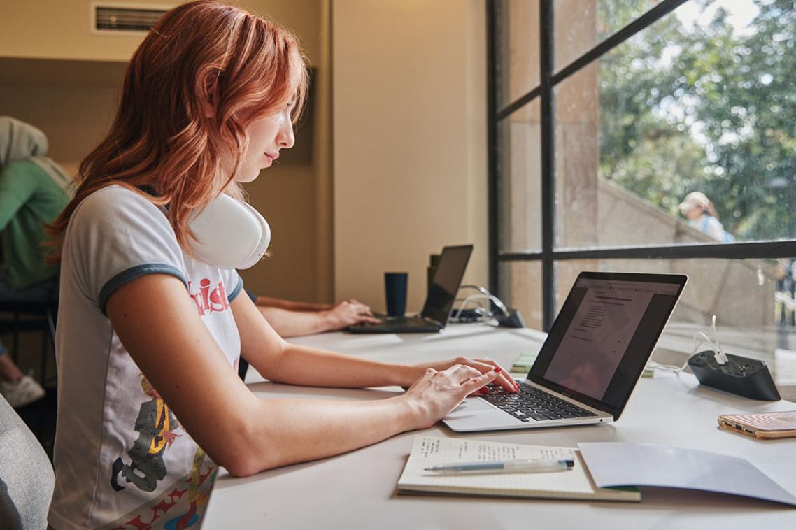 Student seated in front of a window working on a laptop with a notebooks beside them.