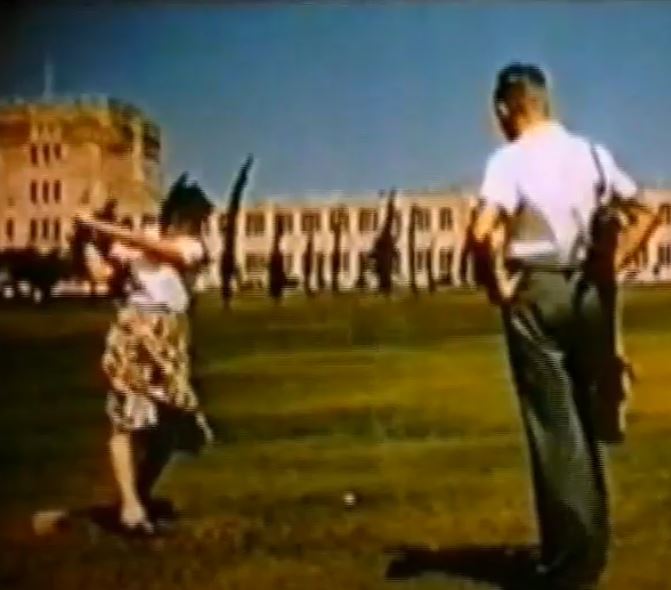 Woman swinging golf club in front of Forgan Smith. A man watches with hands on hips. Green grass, blue cloudless sky.