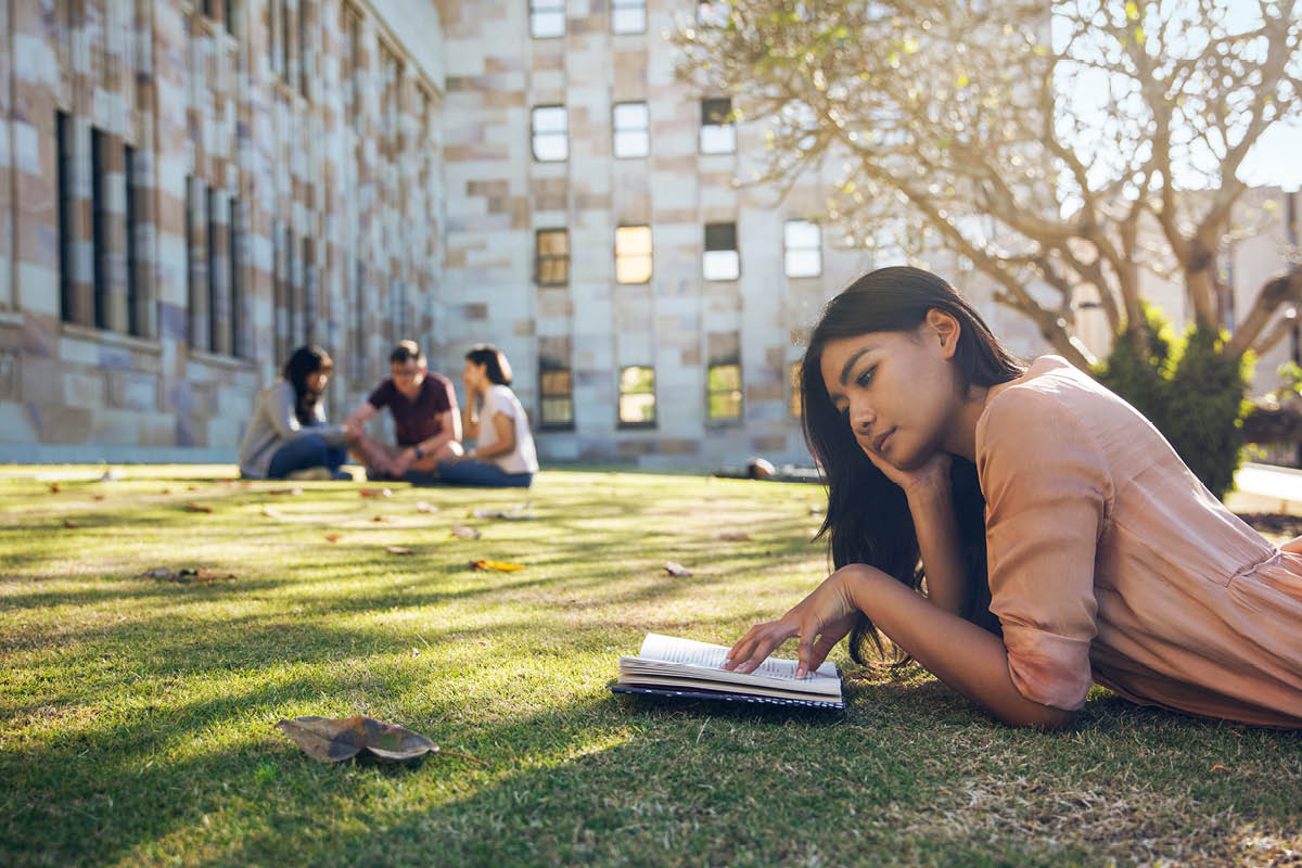 Person reading a book on the grass, near a tree, in the Great Court at UQ. 3 students are sitting together talking nearby.