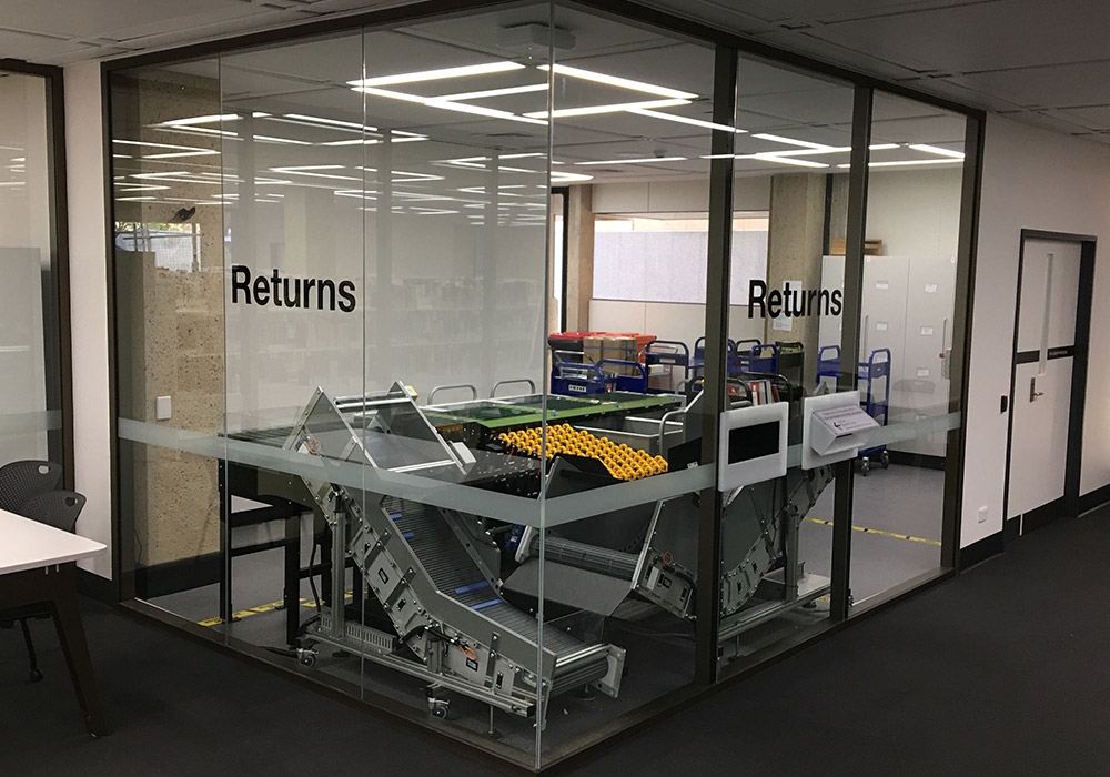 Return chutes in glass wall. Behind the wall is the book machine which returns and sorts books for reshelving. Caption below.