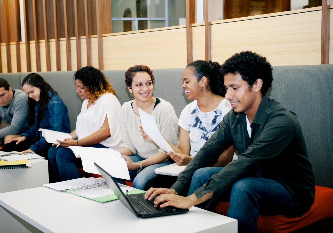 Six students seated on a bench with laptops and books, working and having discussions