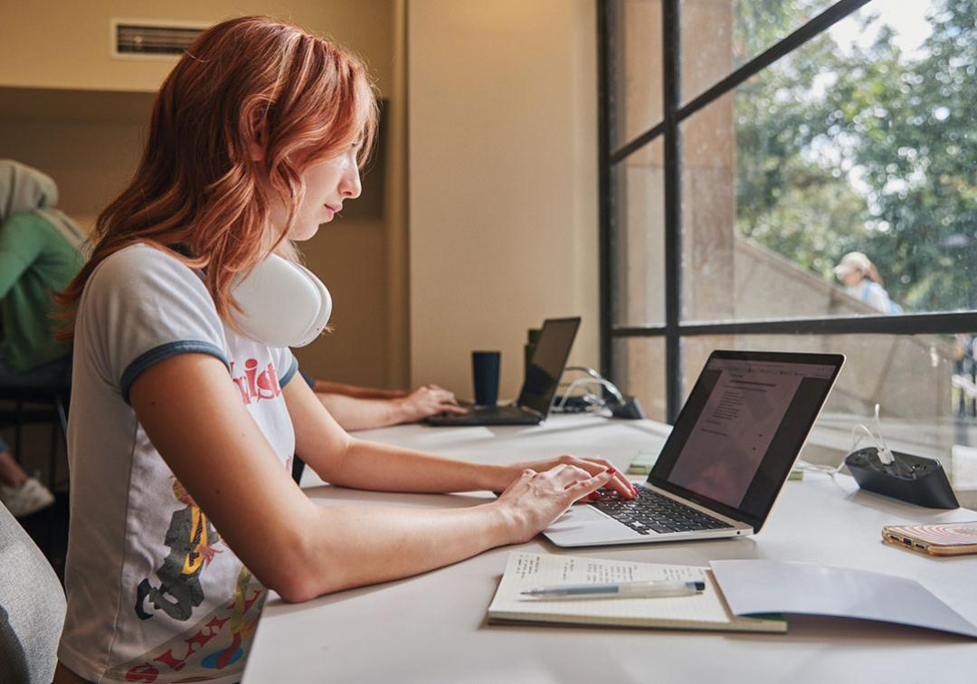 Student seated in front of a window working on a laptop with a notebooks beside them.