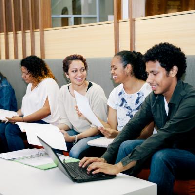 Six students seated on a bench with laptops and books, working and having discussions