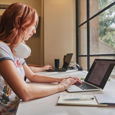 Student seated in front of a window working on a laptop with a notebooks beside them.