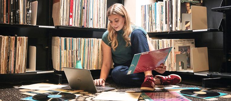 Female student sitting on the floor with records, papers and laptop in Architecture and Music Library.