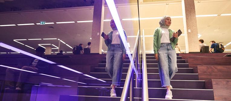 Student walking down the Purple Stairs in Central Library. She is reflected in the purple glass next to her.