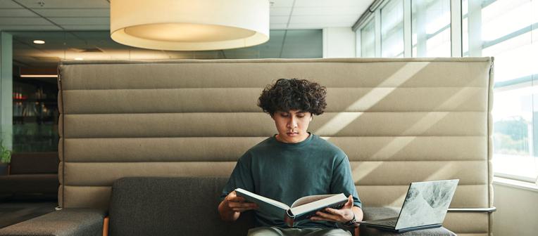 Student with a book and laptop on a lounge in Dutton Park Health Sciences Library.