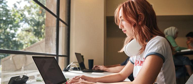 Student facing windows working on a laptop with headphone around neck