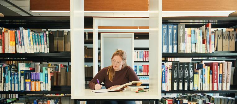 Student studying with open books and taking notes, surrounded by book shelved