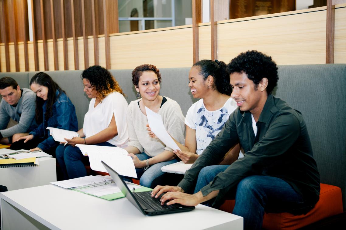 Six students seated on a bench with laptops and books, working and having discussions