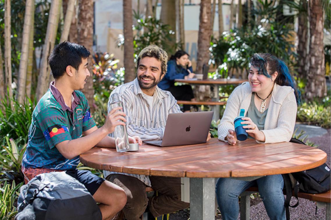 Three people seated at a table outside with a laptop having a discussion