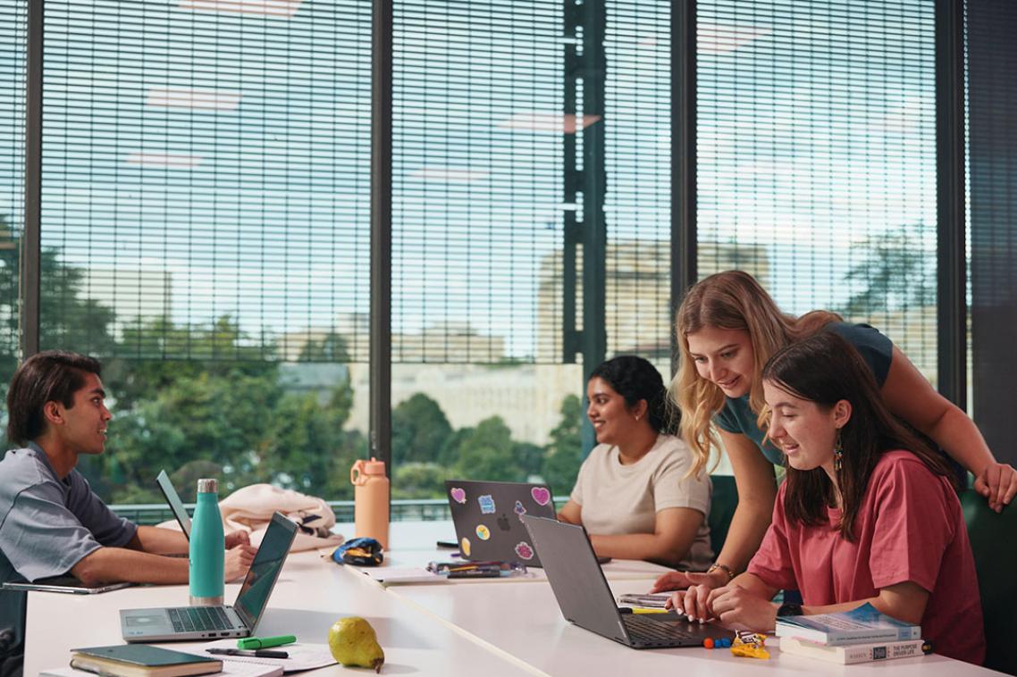 Fours students at a desk with one standing, all looking at laptops with a large window in the background.