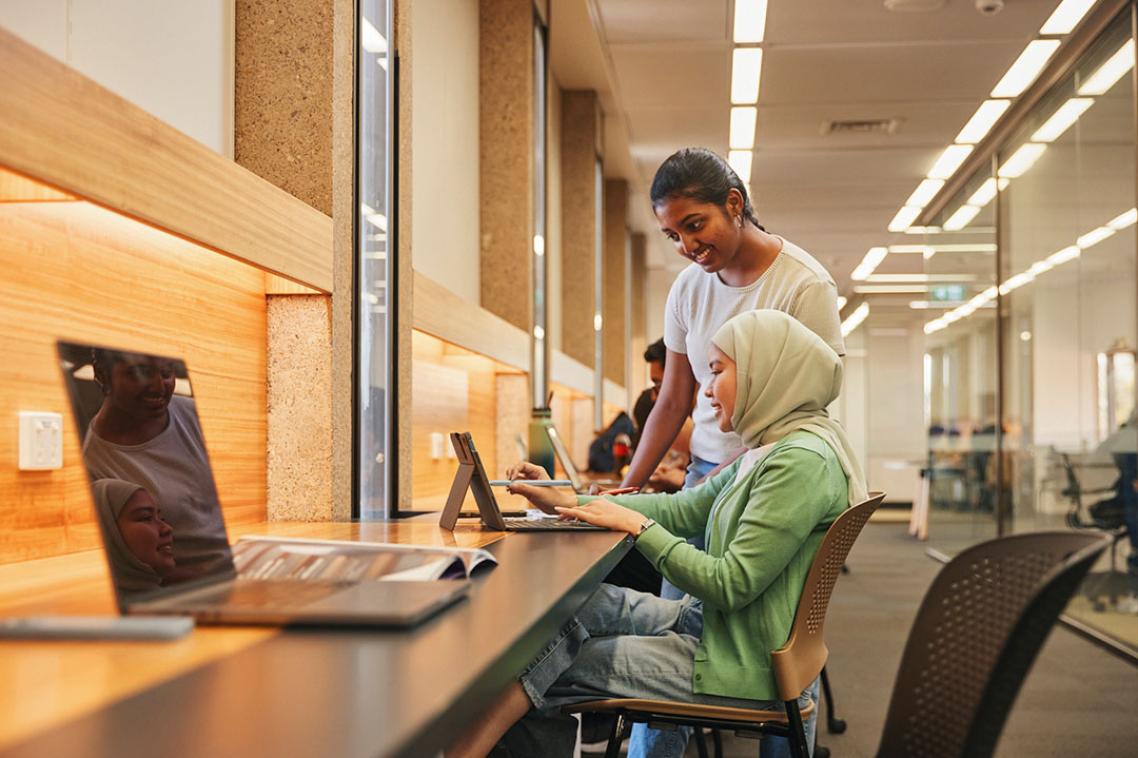 Two students, one sitting and one standing, looking at a laptop on a desk.