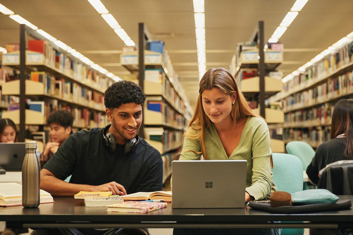 Two students at a desk looking at a laptop with book shelves in background.