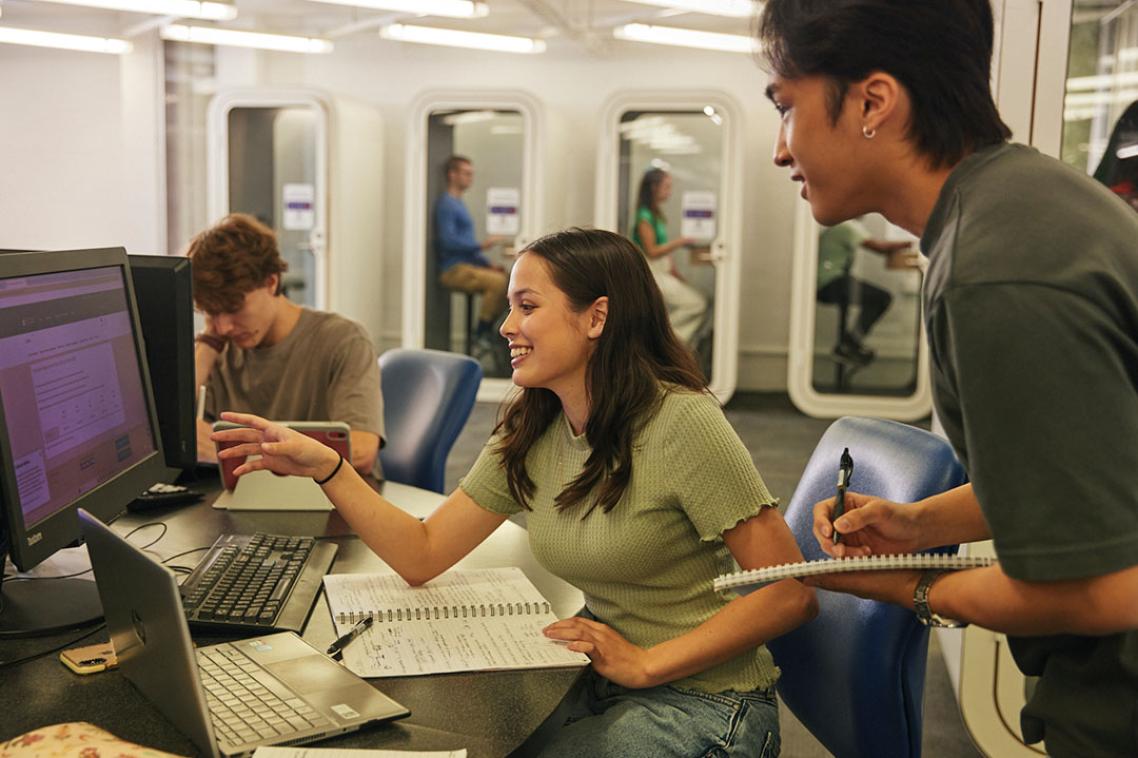 Two students looking at computer, one standing, with open books and taking notes