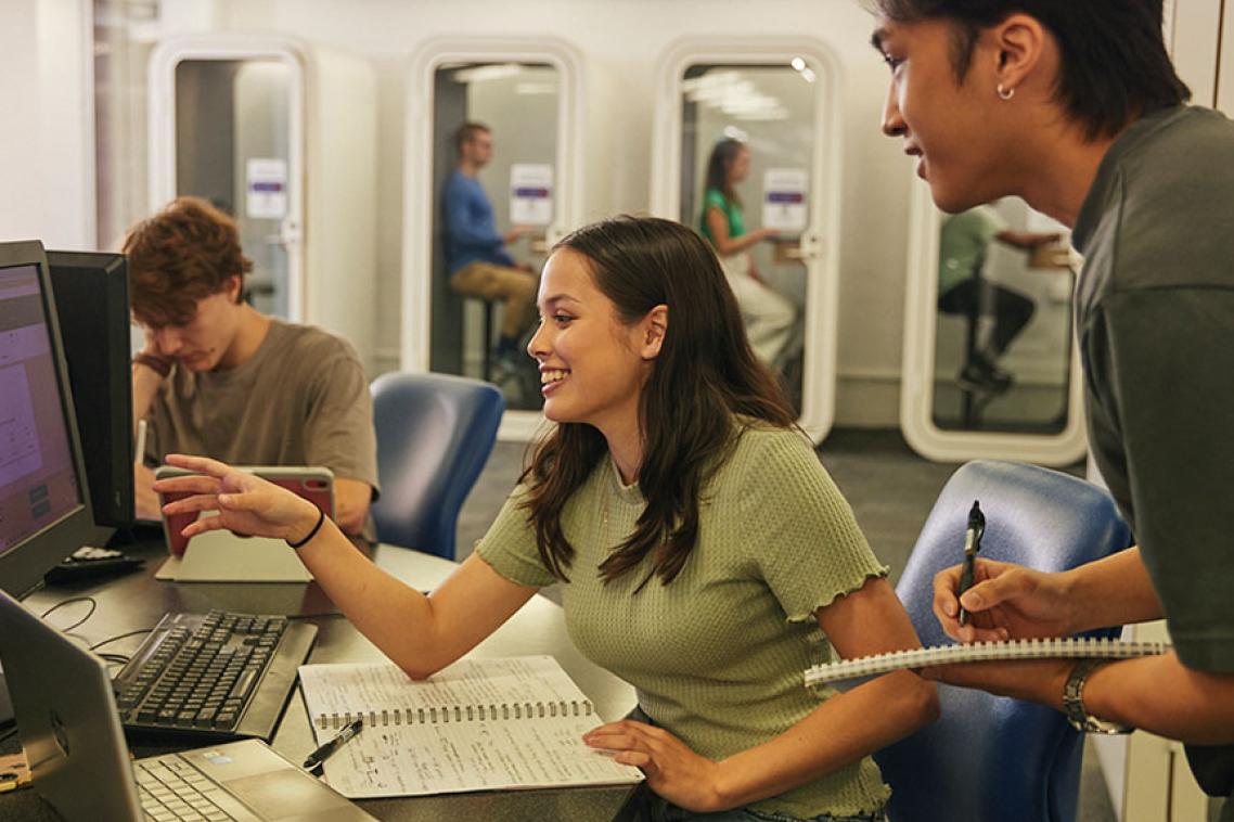 Two students looking at a computer, one standing, open books on desk and one student taking notes.